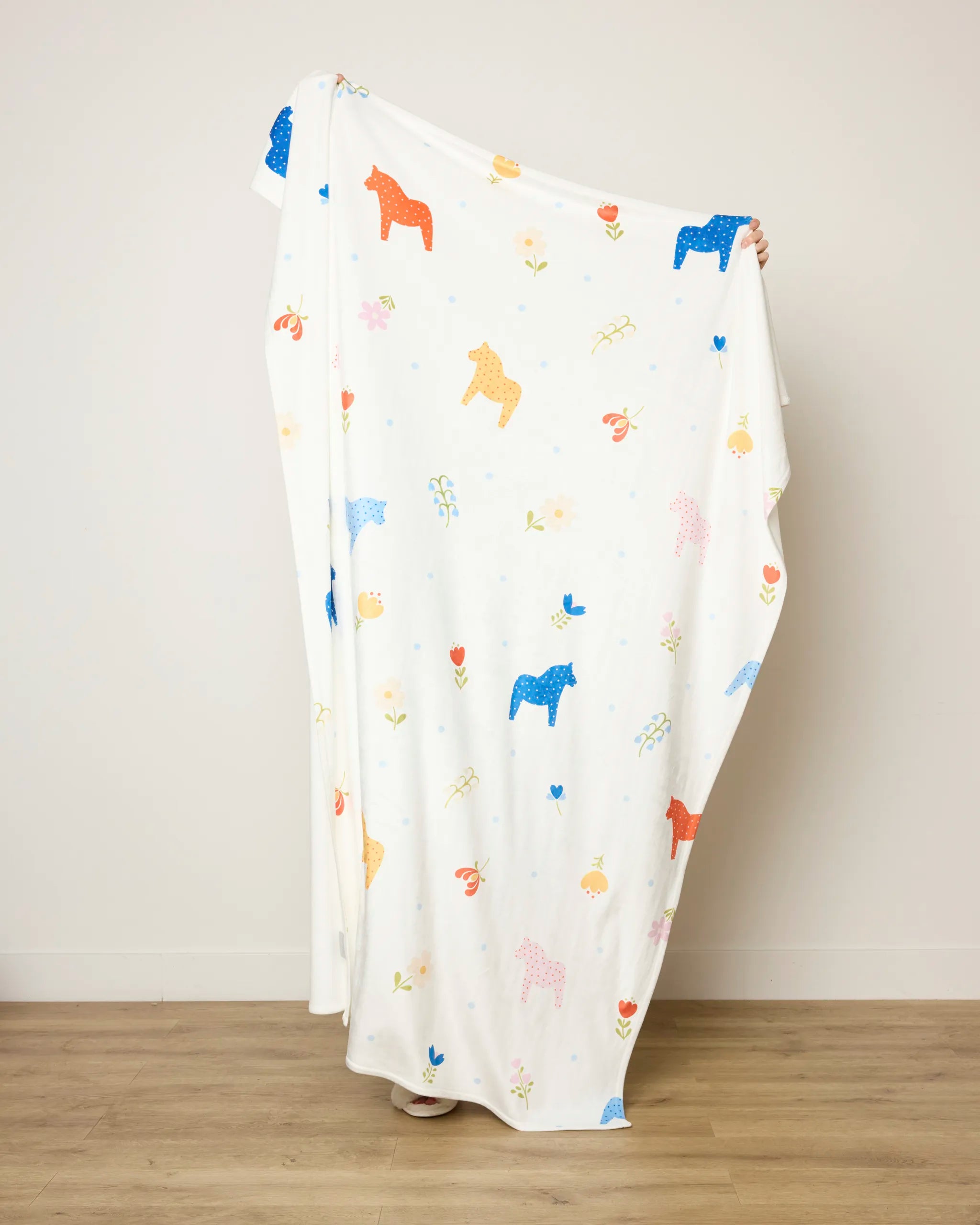 A person holds up the Sara Mari Dala Garden Floral throw blanket, featuring colorful horse, flower, and leaf patterns. The soft blanket covers most of their face and body while they stand against a plain wall and wooden floor.