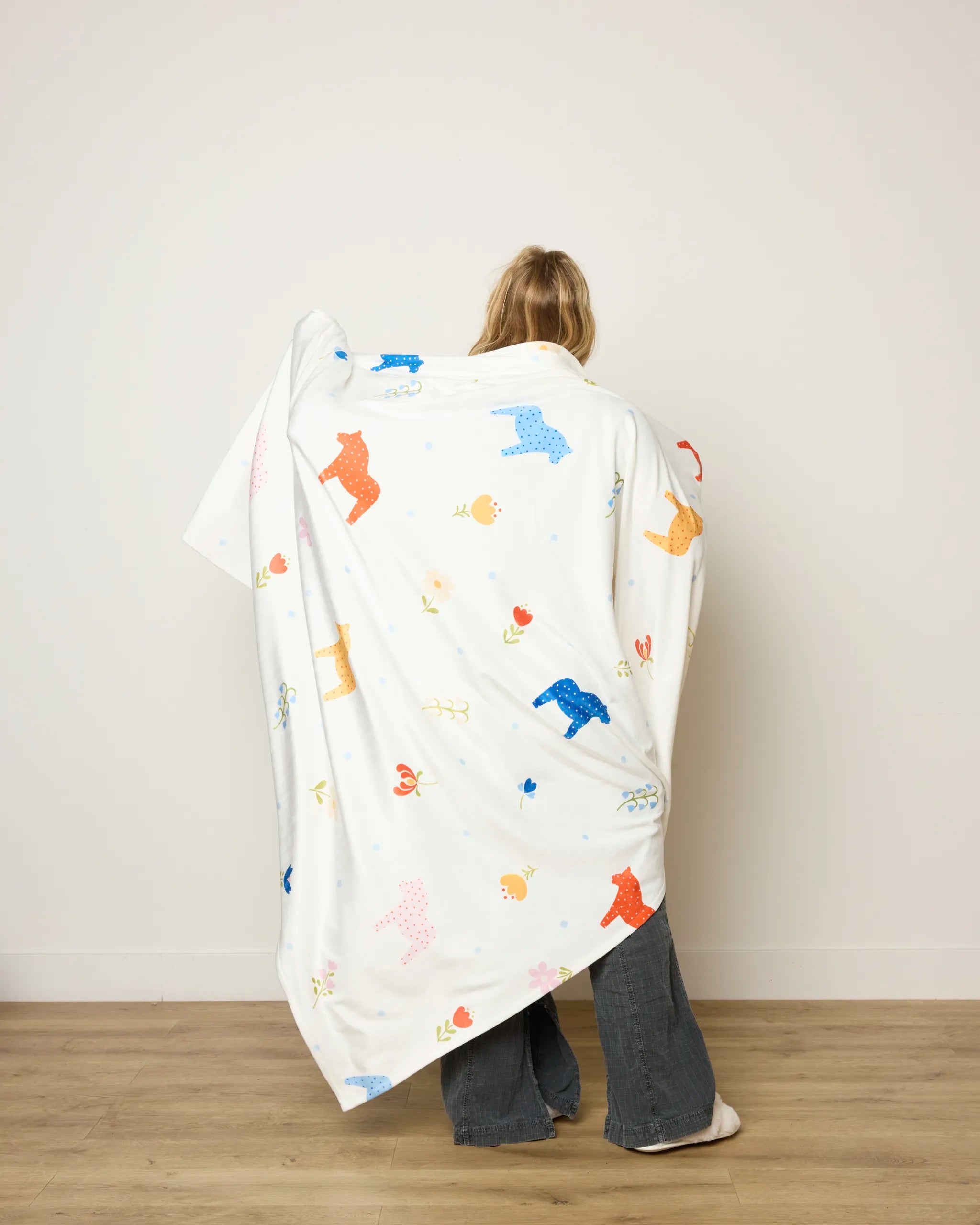 A person stands with their back to the camera, wrapped in the Sara Mari "Dala Garden Floral" throw blanket, featuring colorful animal and flower prints. They wear gray pants and stand on a light wooden floor against a plain wall.