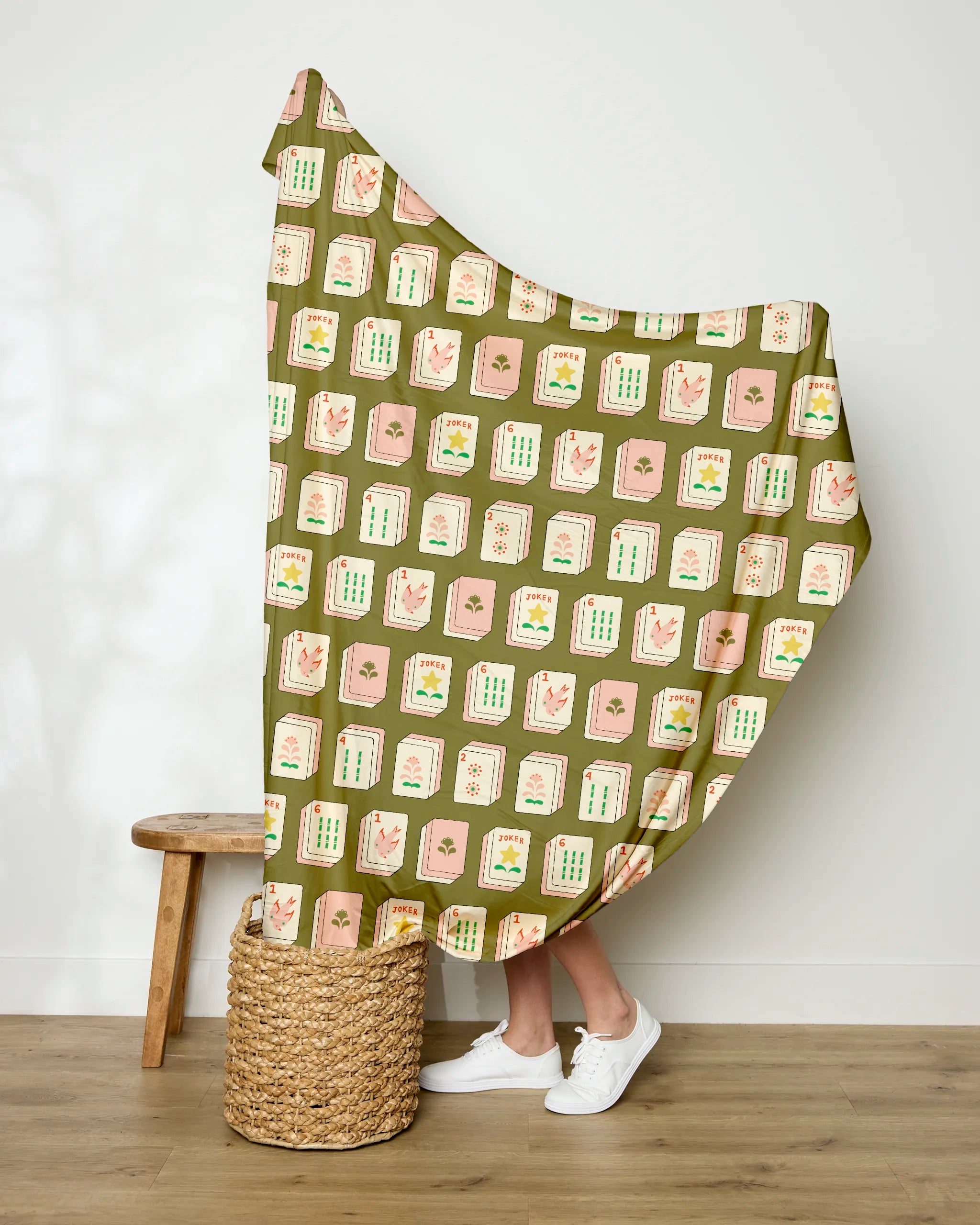 A person holds up the Geometry "Flower Tiles Green" soft throw blanket, featuring mahjong tile designs, while standing in a woven basket. Only their legs and white sneakers are visible. A wooden stool sits nearby against a white wall.
