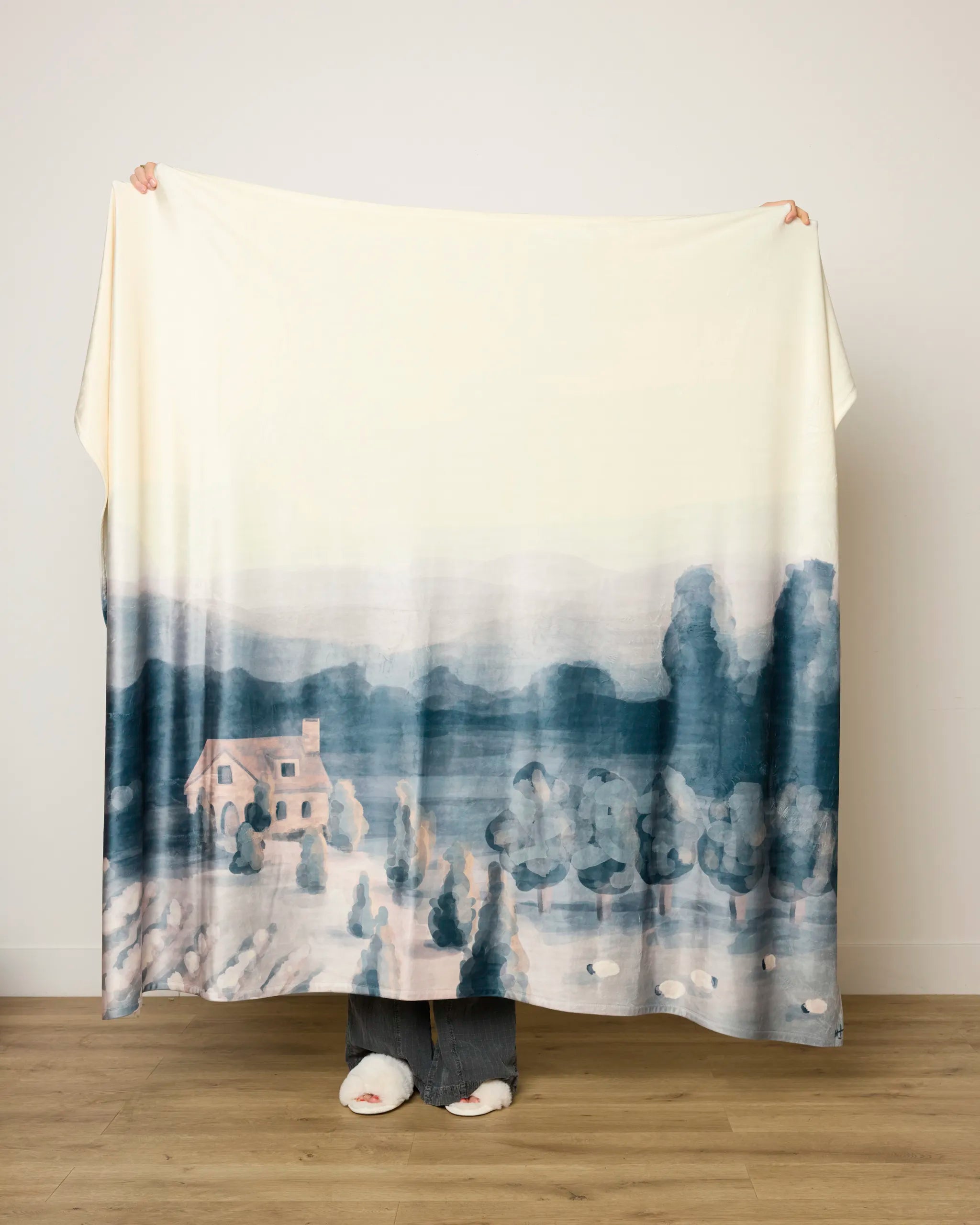 A person holds up the "French Cottage" butter blanket by Hannah Langford, featuring a watercolor landscape of a house, trees, and mountains in blue and muted pink tones. Only their hands, feet, and jeans are visible.