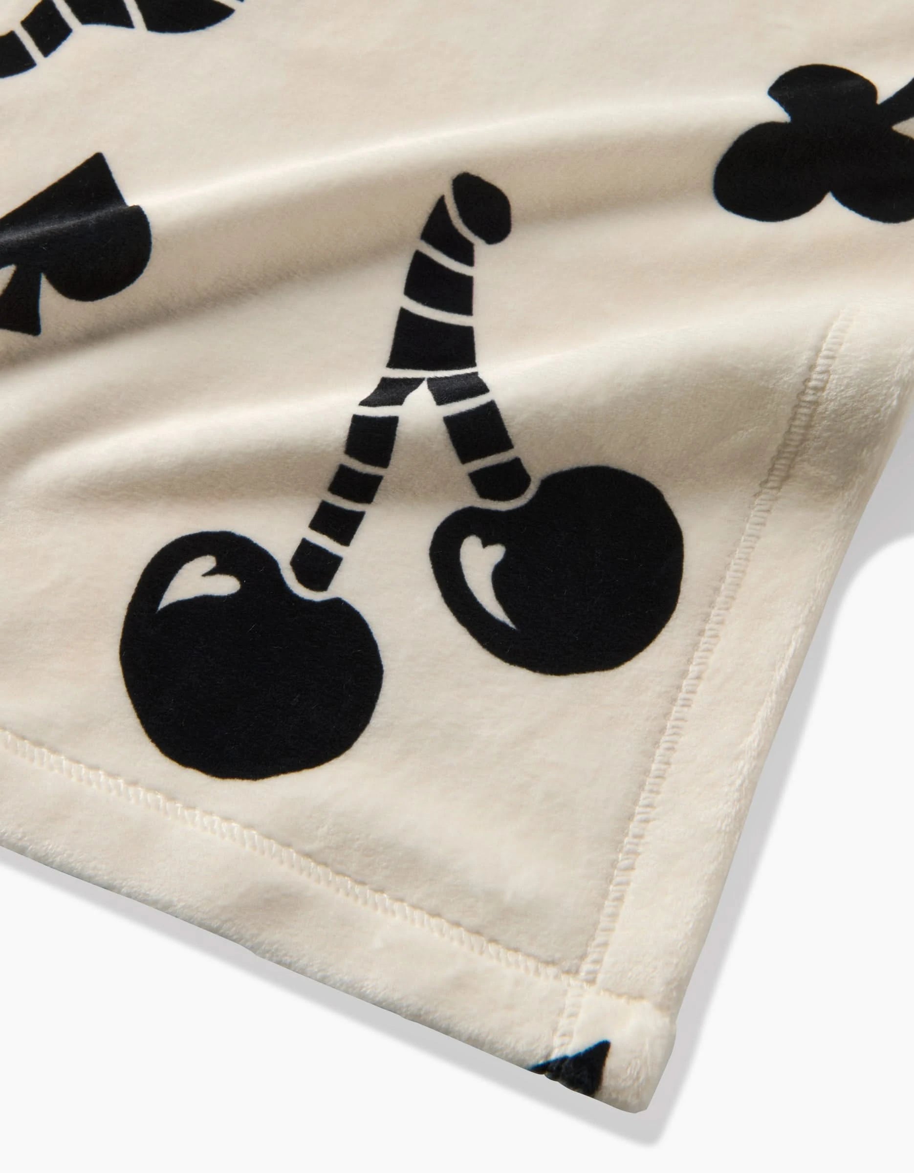 A close-up of the Heart Locket blanket by Geometry, cream-colored and made from recycled materials, with black graphic prints of cherries and playing card suits, stitched edges, and a silky-soft feel.