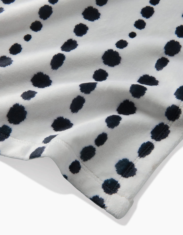 The Geometry Ink Dots Butter Blanket is a cozy, eco-friendly white fleece made from recycled materials, featuring large and small black dots arranged diagonally and a partially folded edge that highlights its inviting warmth.