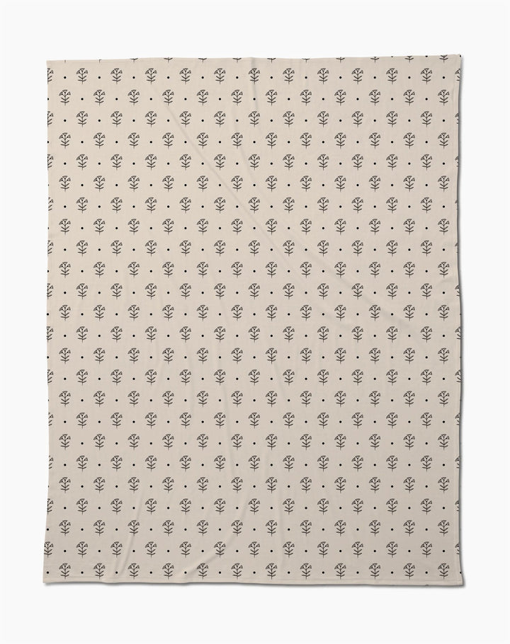 The Inked Bloom by Cole Strem is an eco-friendly, beige blanket with a simple, repeating black floral motif. Made from recycled materials, it appears soft with subtle natural wrinkles.