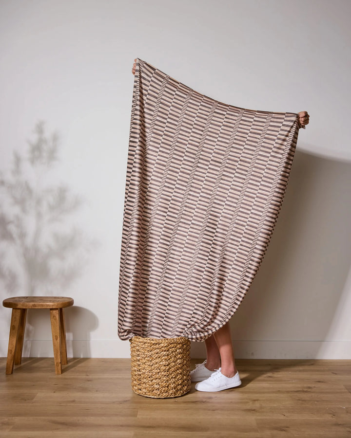A person stands on a woven basket, holding up the Geometry Light Ridge butter blanket, which covers their upper body. Only their legs in white sneakers show, with a wooden stool and plant shadow in the background.