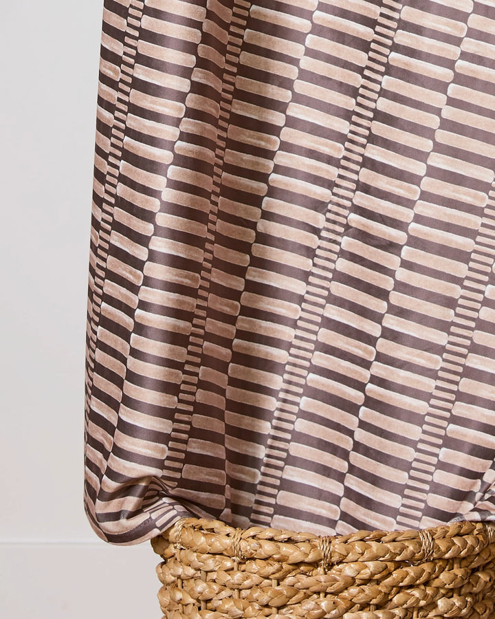 A close-up of Geometrys Light Ridge recycled blanket, featuring a brown and beige geometric pattern, draped over a woven natural-colored basket against a neutral background.