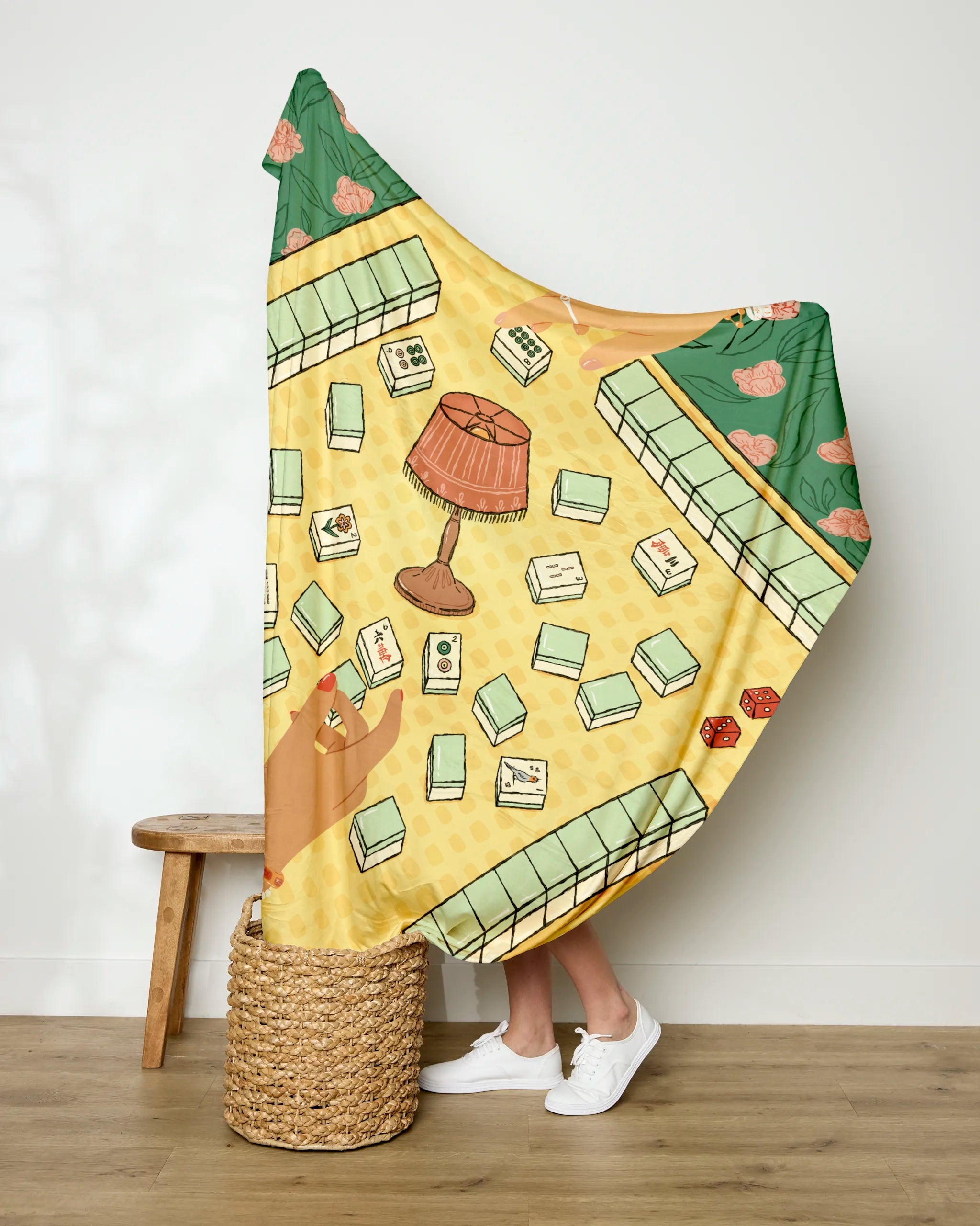 A person stands against a white wall holding up the "Lucky Hand" throw blanket by Geometry, which features a Mahjong-themed illustration with scattered tiles, a central lamp, two red dice, and hands mid-game over a green floral border. A wooden stool and basket sit nearby.