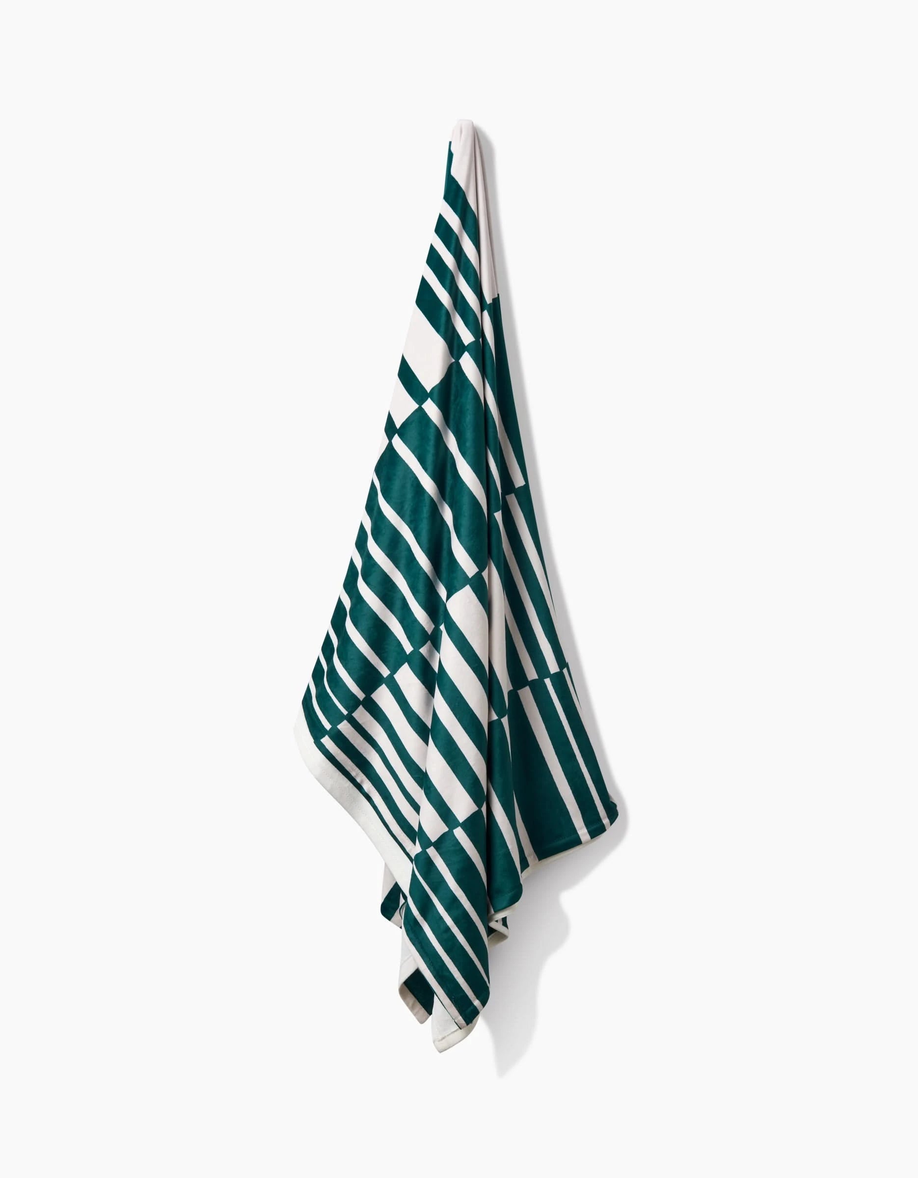 A Verdelia towel by Geometry, featuring dark green and white diagonal and horizontal stripes, hangs on a wall, offering silky softness against a plain white background.