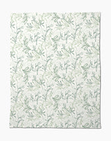 The Organic Foliage Butter Blanket by Kris Ruff is white, silky soft, and features a subtle repeating pattern of green leafy branches and vines for a delicate botanical look.