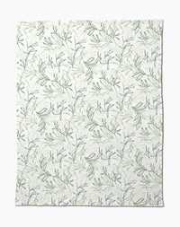 The Organic Foliage Butter Blanket by Kris Ruff is white, silky soft, and features a subtle repeating pattern of green leafy branches and vines for a delicate botanical look.