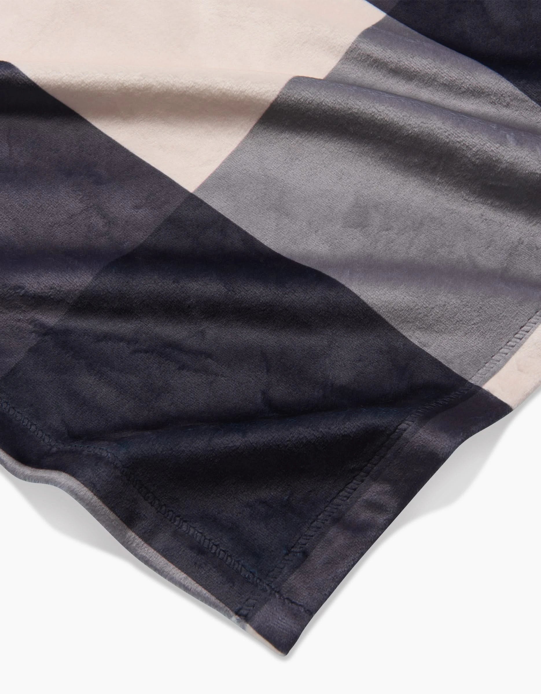 A close-up of Geometry’s Snowy Gingham blanket, made from recycled materials, features large overlapping rectangular blocks in beige, gray, and dark blue, showcasing its silky softness and detailed edge stitching.