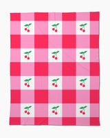 The Sweet Cherry Gingham throw blanket by Geometry features a soft fabric with a red and pink checkered pattern, accented by charming cherry illustrations with green leaves in the center of select squares.