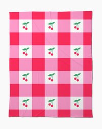 The Sweet Cherry Gingham throw blanket by Geometry features a soft fabric with a red and pink checkered pattern, accented by charming cherry illustrations with green leaves in the center of select squares.