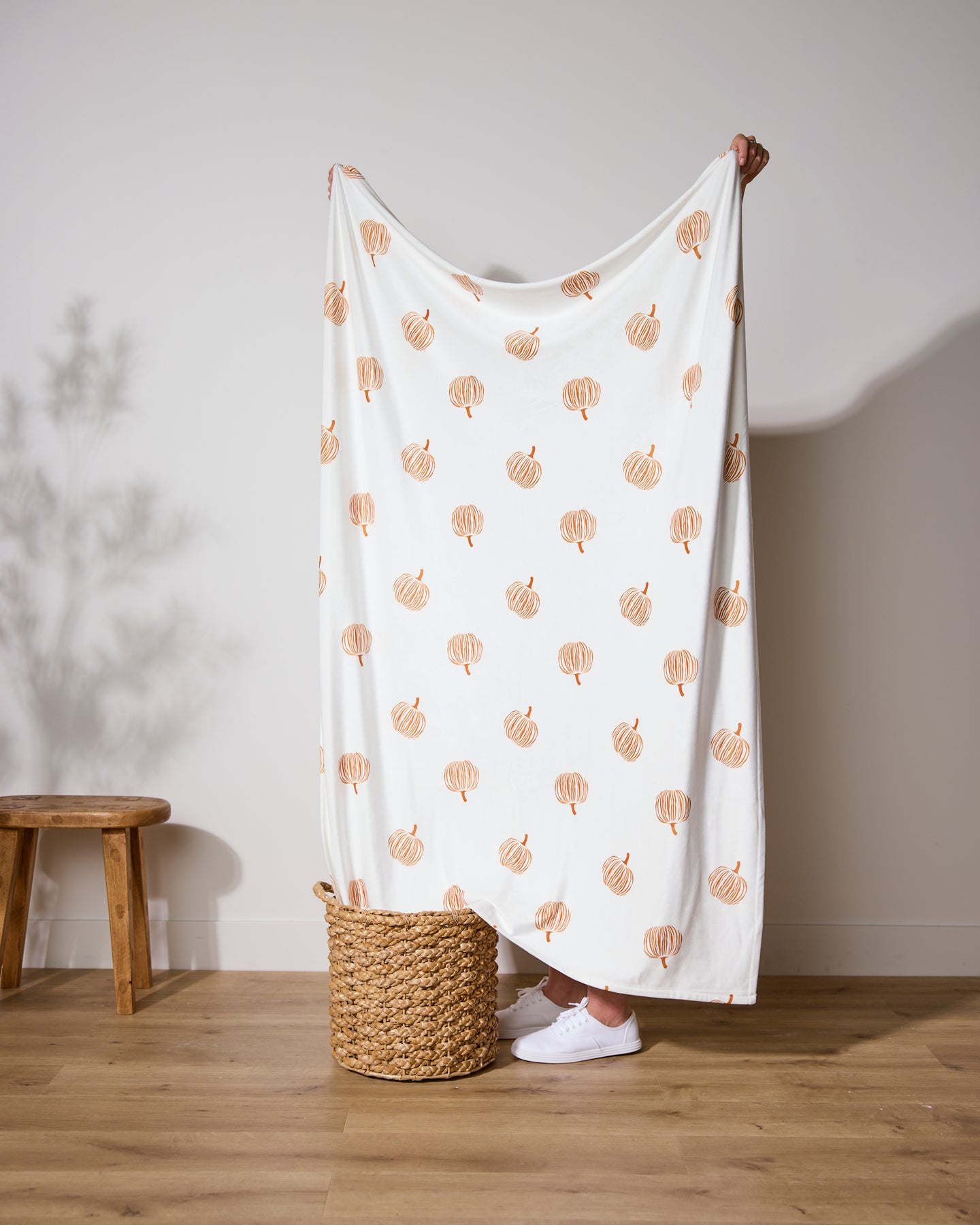 A person holds up the Geometry “Tales From the Patch” Butter Blanket, featuring orange pumpkin illustrations on white, hiding behind its soft fabric. The throw drapes into a woven basket next to a wooden stool on light wood flooring.