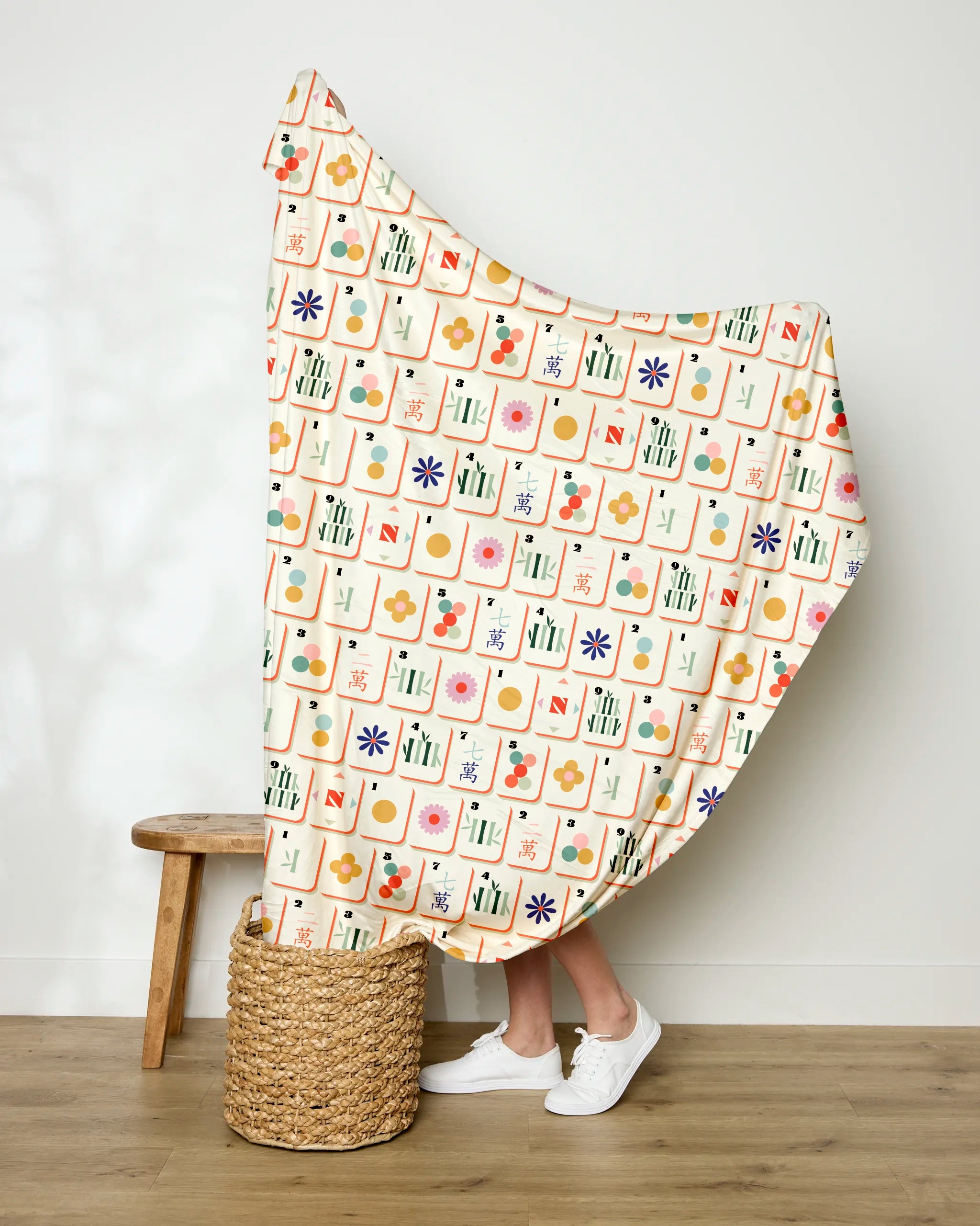 A person stands behind a colorful patterned Tile Toss blanket by Geometry, holding it up. Only their lower legs and white sneakers are visible. The scene includes a wooden stool and a woven basket on a hardwood floor.