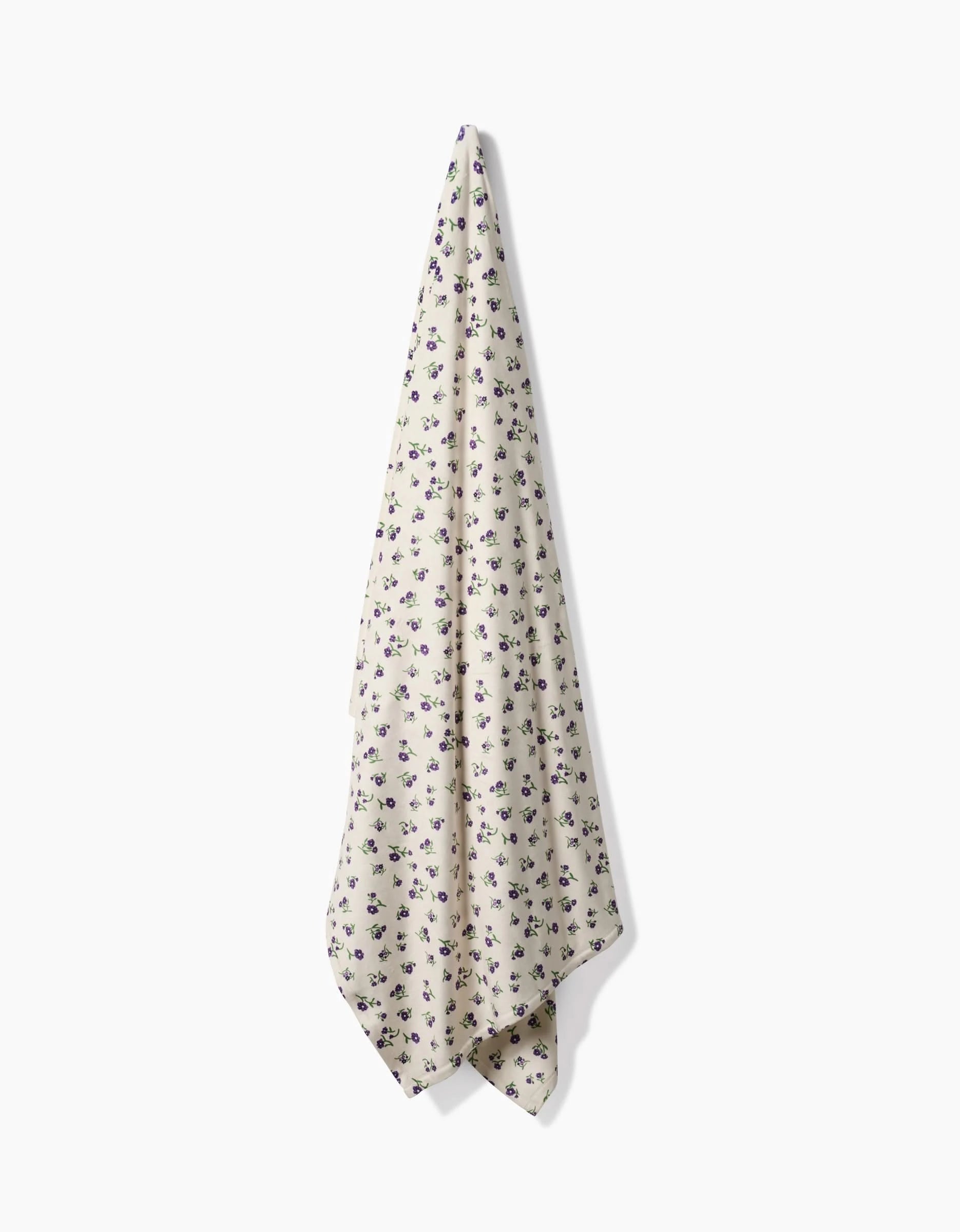 A cream-colored Geometry Violet Sound Butter Blanket, made from recycled materials and featuring a small purple and green floral pattern, hangs from a point against a white background.