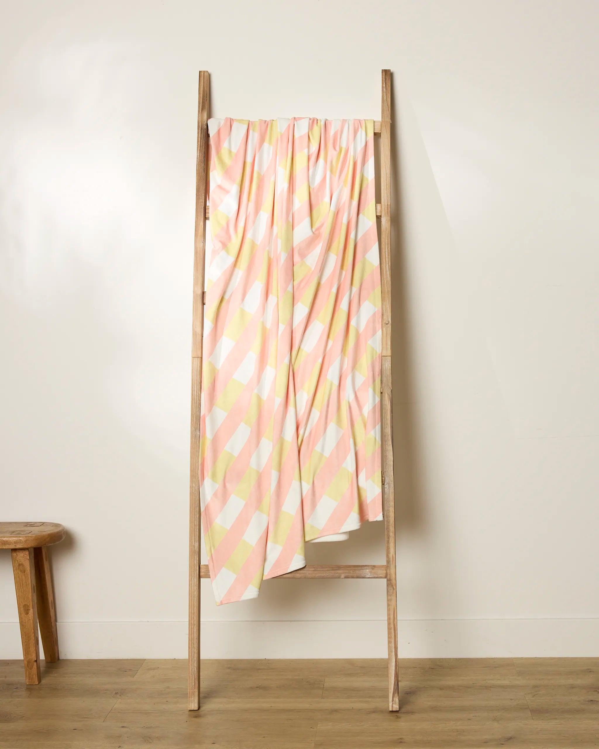 A wooden ladder leans against a white wall, holding Geometry’s Apricot Glow, a pink and white checkered throw blanket made from recycled materials. A small wooden stool rests nearby on the light wood floor.