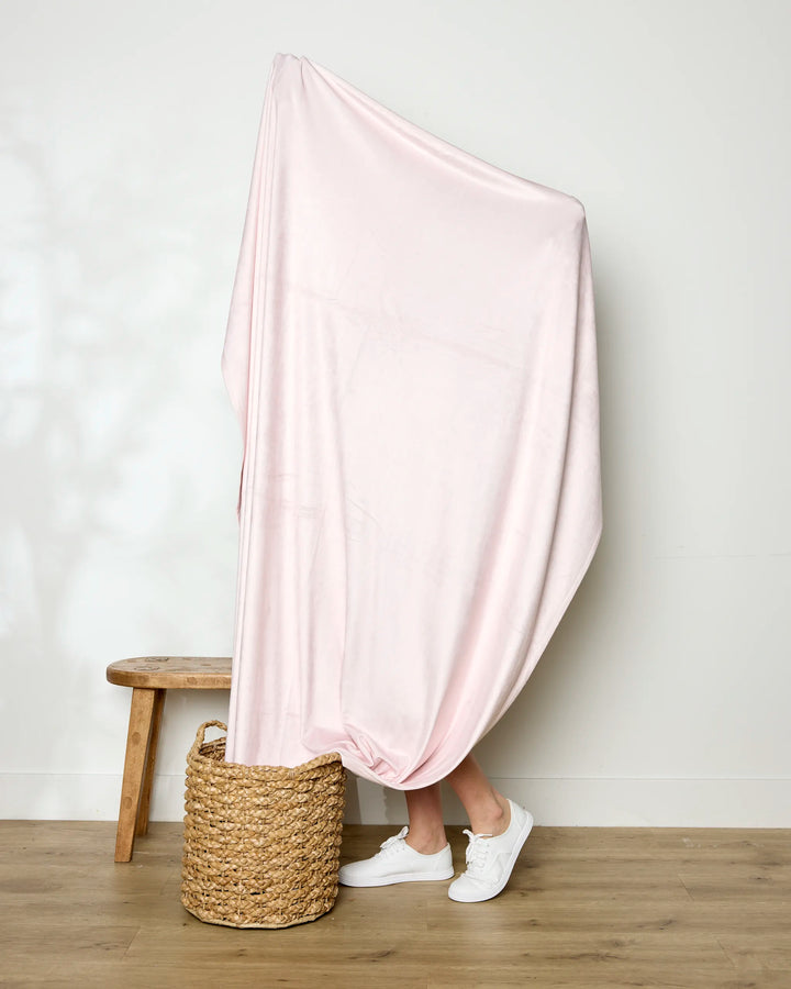 A person stands behind a large, light pink GEOMETRY Berry Shortcake blanket that completely covers them, revealing only their lower legs and white sneakers. Nearby are a woven basket and wooden bench against a minimalist backdrop.