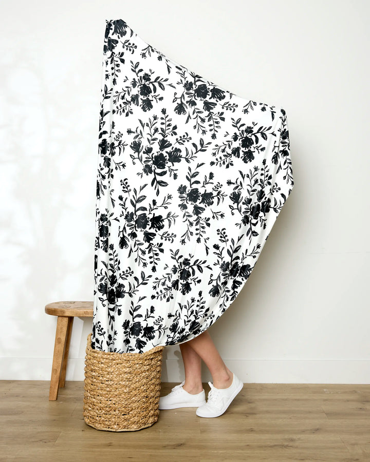 A person holds up the large white Danelys Sidron Botanical Silhouettes Charcoal blanket, featuring a black floral pattern. Only their lower legs and white sneakers are visible near a woven basket and wooden stool on a wood floor.