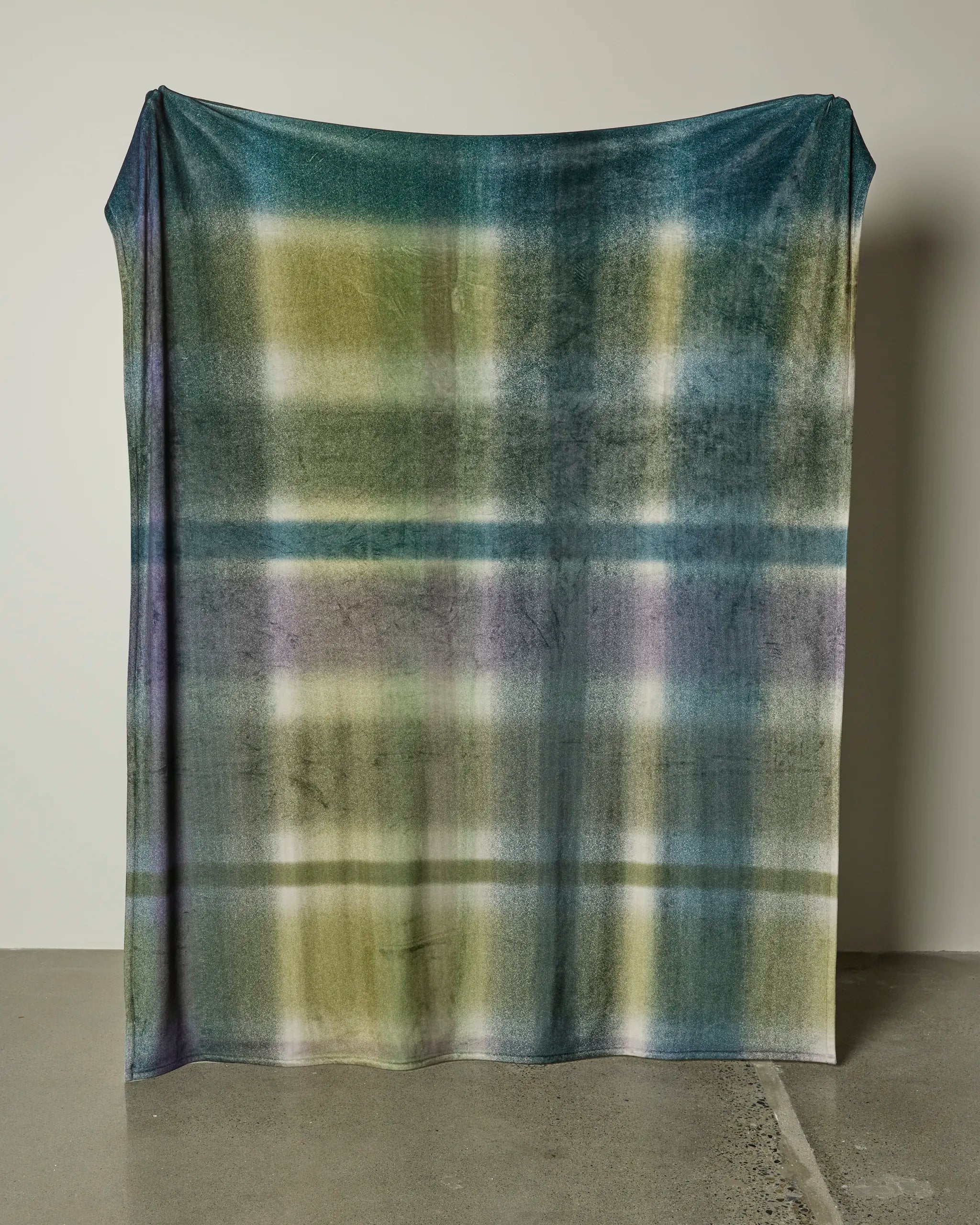 The Geometry Bracken throw blanket, featuring a soft, oversized design and a blurred plaid pattern in green, blue, yellow, and purple, hangs vertically against a light wall and drapes onto a polished concrete floor.