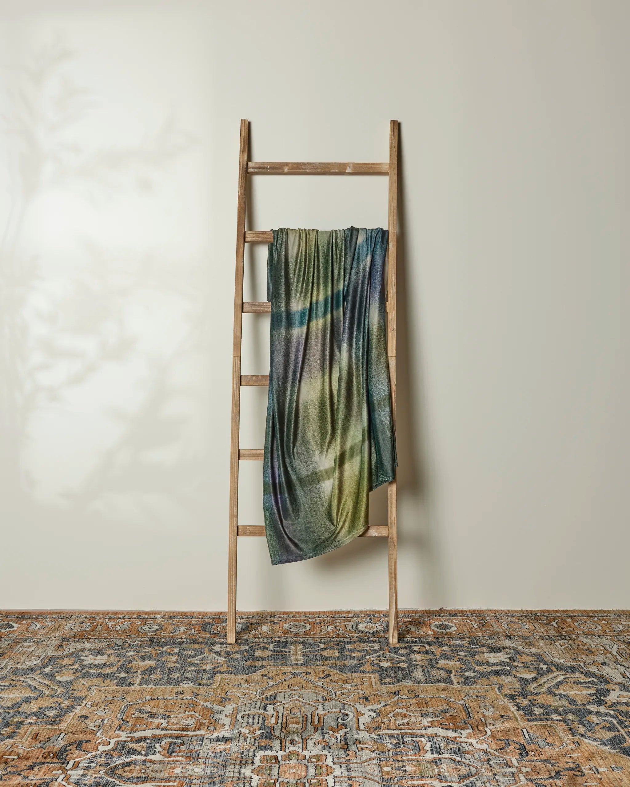 A Bracken recycled blanket by Geometry is draped over a wooden ladder against a light wall. Below, an ornate blue and brown rug covers the floor, while gentle shadows play across the scene.