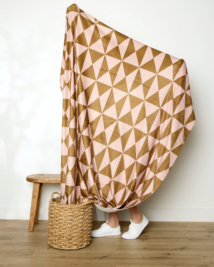 A person stands behind the Geometry "Bronze" recycled blanket, draped from a basket to above their head, concealing their body except for white sneakers. A wooden bench sits nearby on the wood floor.