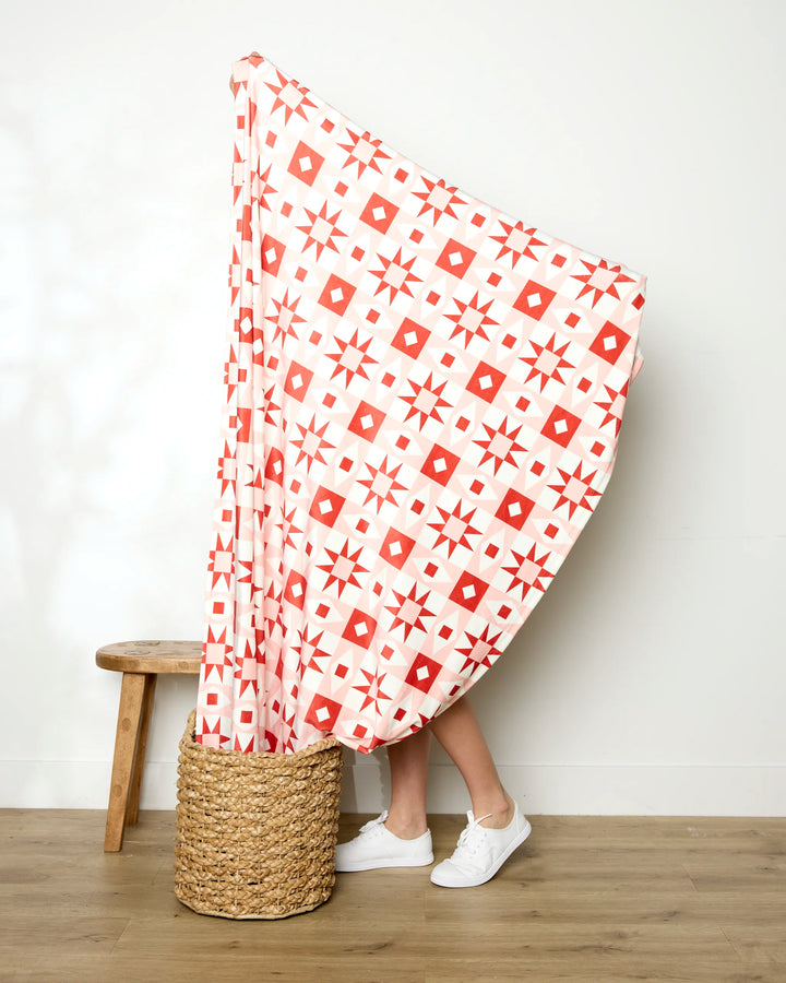A person stands behind the Chalet red and white geometric soft throw blanket by Charlie Rowan Designs, with only their legs in white sneakers visible beside a woven basket and wooden stool on a wooden floor.