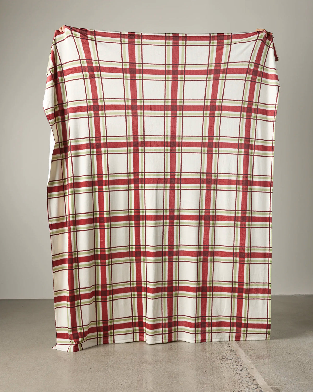 A large sheet, resembling Geometry’s Cozy Cabin red and white plaid Butter Blanket, is draped over an unknown object, standing against a plain light gray wall on a concrete floor.