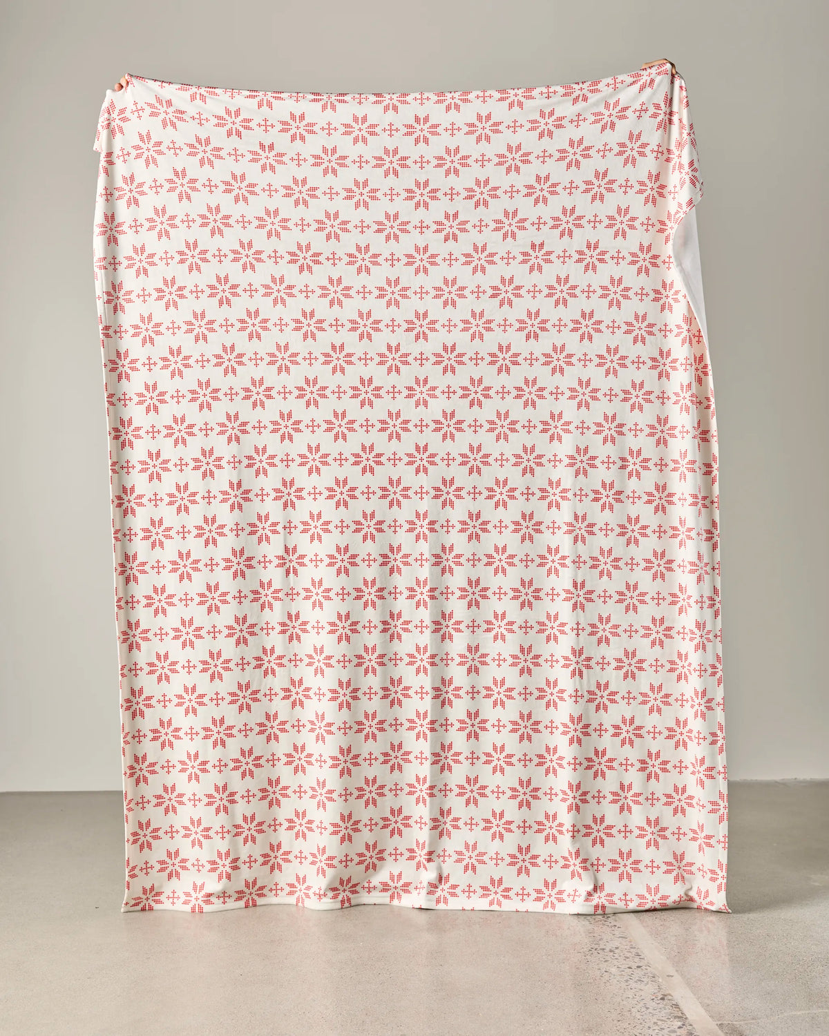 A large rectangular “Cozy Nap” blanket by Geometry, adorned with a pattern of pink star-shaped flowers, is held upright, concealing whatever is behind it. The background consists of a plain light-colored wall and floor.