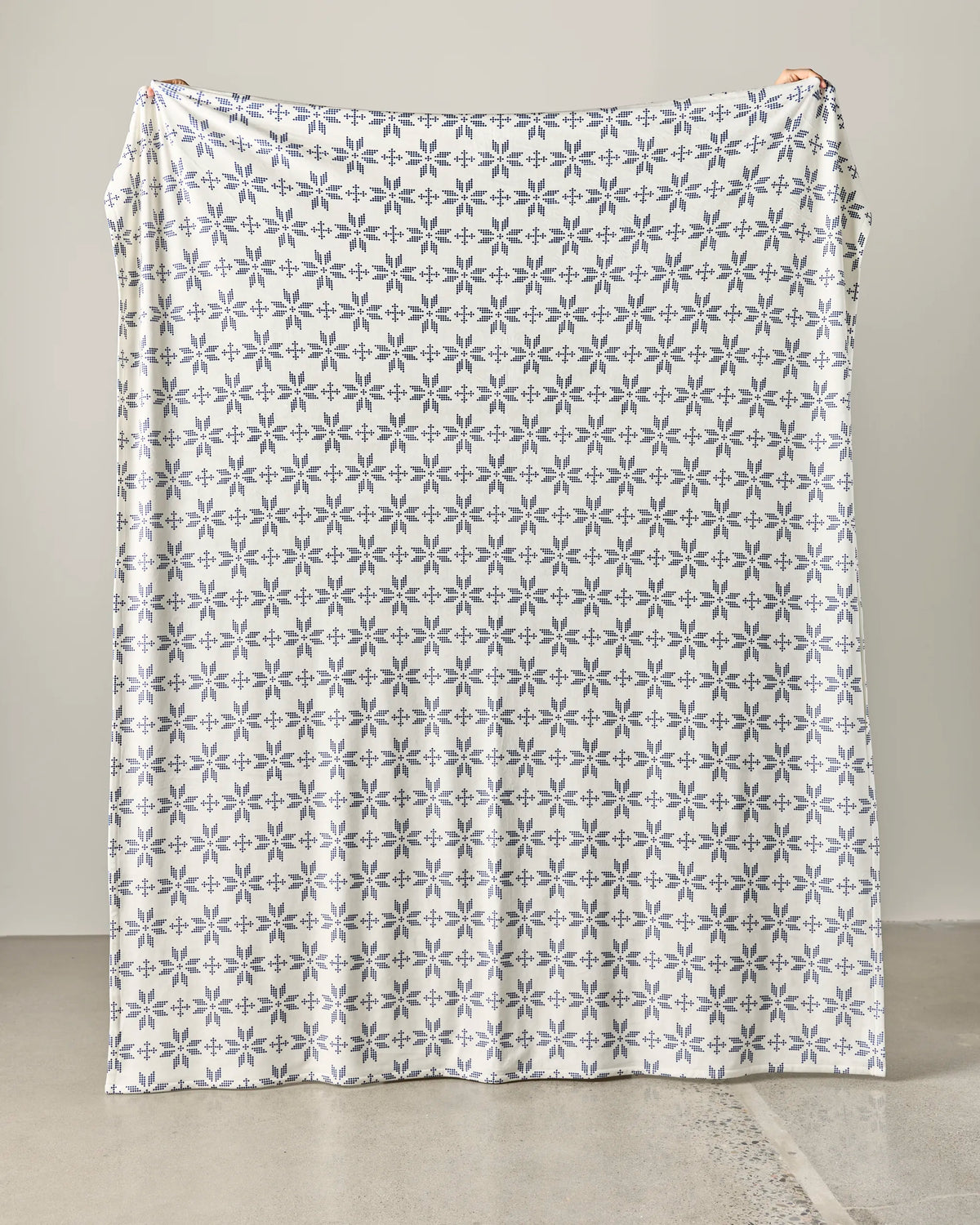 A person holds up the Geometry “Cozy Nap” blanket—an oversized white throw featuring a repeating dark blue floral and geometric design—against a plain light background, standing on a gray floor with only their hands visible at the top corners.