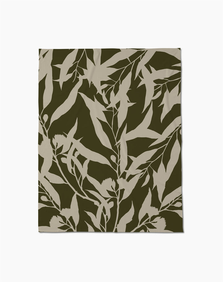 The Geometry Fable Butter Blanket features a botanical pattern of beige leafy branches on an olive green background—perfect for cozy evenings.