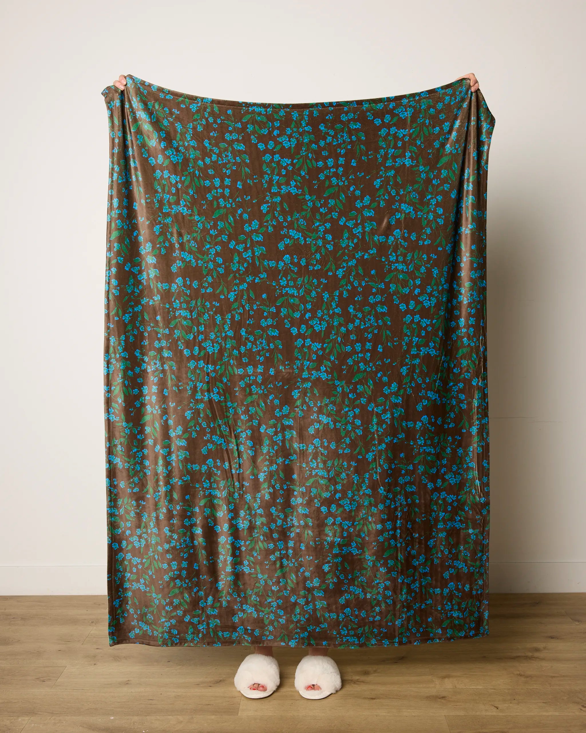 A person stands on a wooden floor, wrapped in the Floralie blanket by Geometry, its blue floral patterns and brown butter color concealing them except for their feet in white slippers.