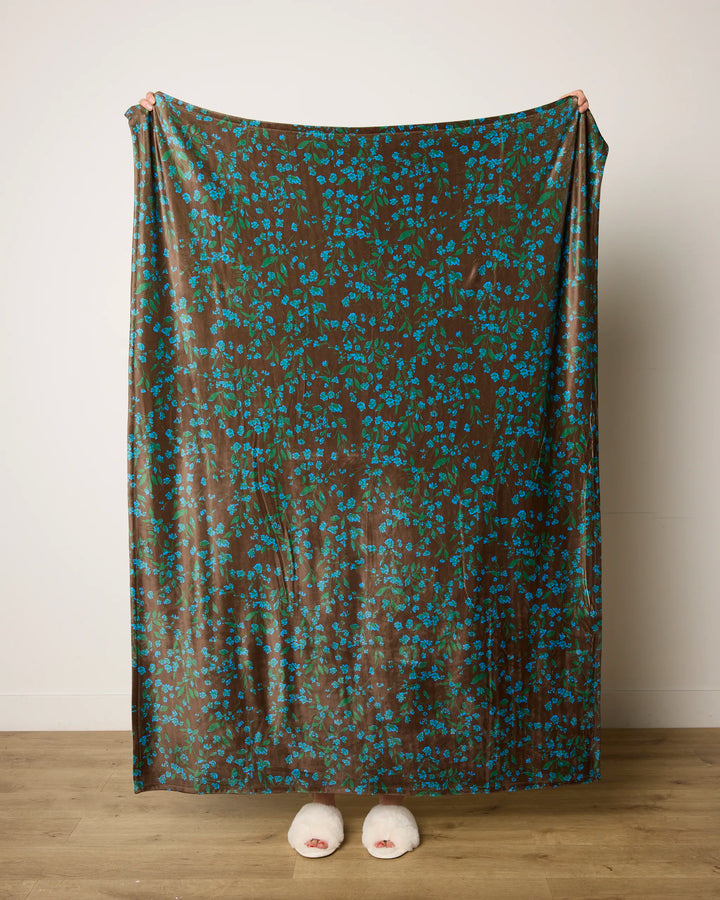 A person stands on a wooden floor, wrapped in the Floralie blanket by Geometry, its blue floral patterns and brown butter color concealing them except for their feet in white slippers.