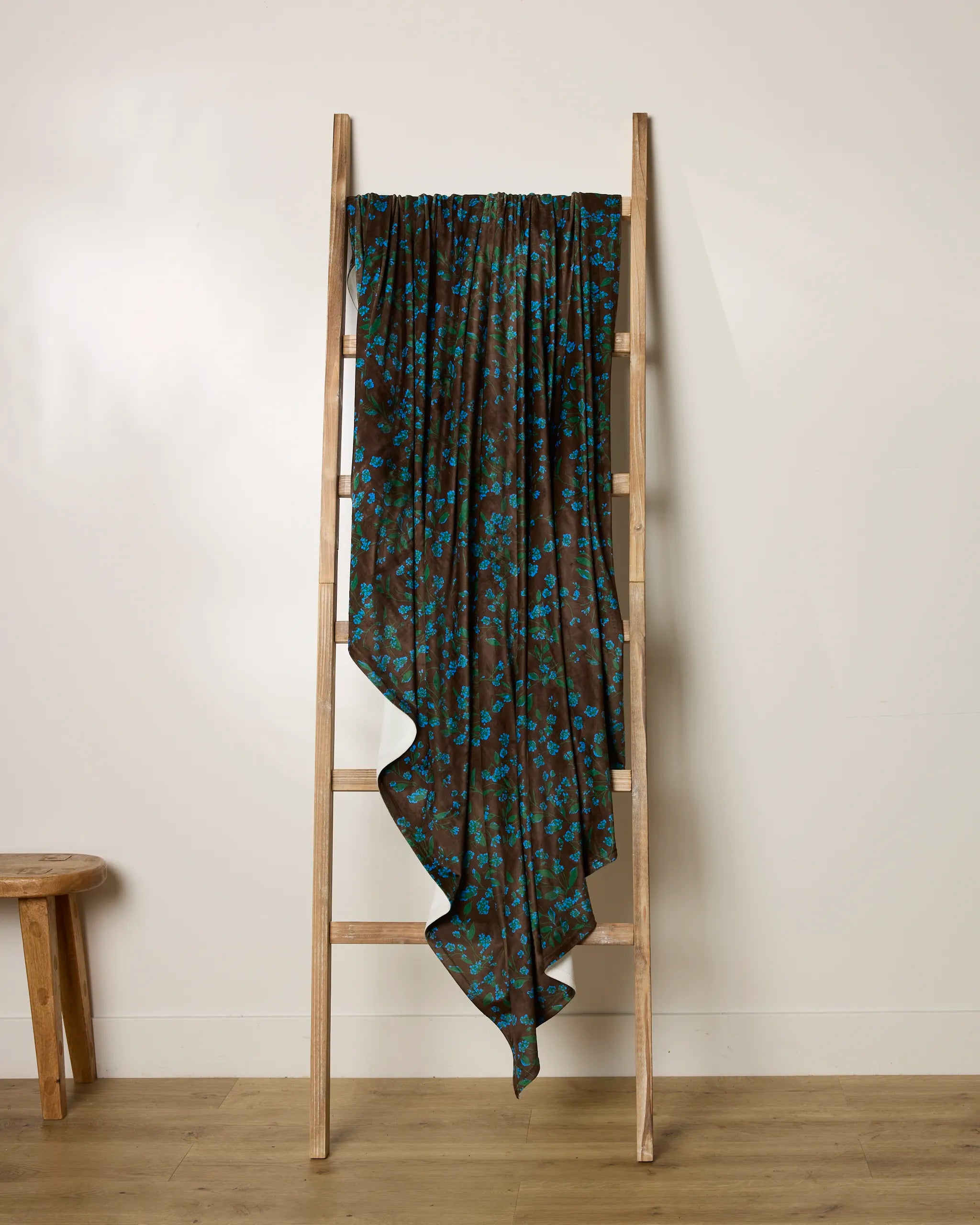 The Floralie scarf by Geometry, featuring a blue floral pattern on brown, drapes over a light wooden ladder set against an off-white wall with a wooden floor. A small wooden stool appears partially on the left side of the image.