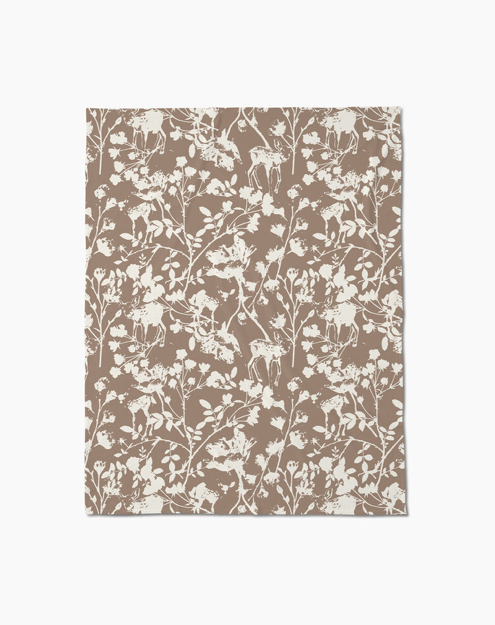 The Forestry throw blanket by Geometry is a soft brown blanket featuring an intricate white forest-themed pattern with deer, birds, flowers, and branches on a plain background.