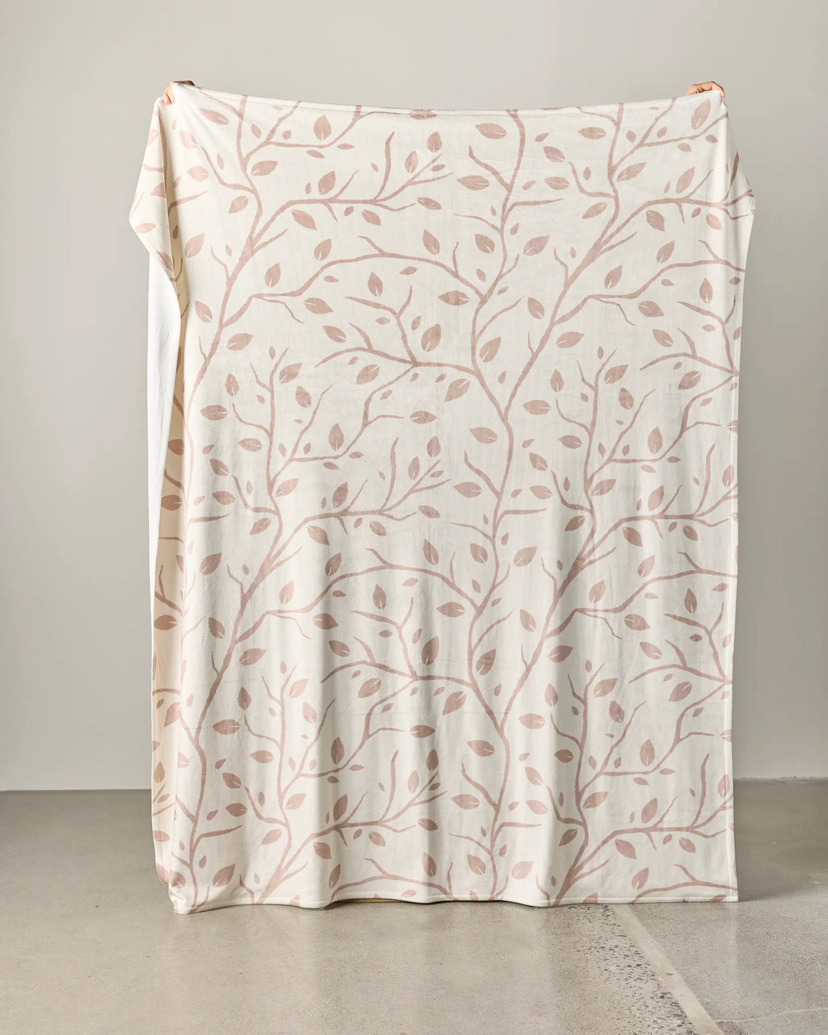 A large Frosted Birch Butter Blanket by Geometry, featuring a cream base and a light brown leafy branch pattern, is held up against a plain light-colored wall, partially resting on the smooth concrete floor. This soft throw blanket is made from recycled materials, providing both comfort and sustainability.