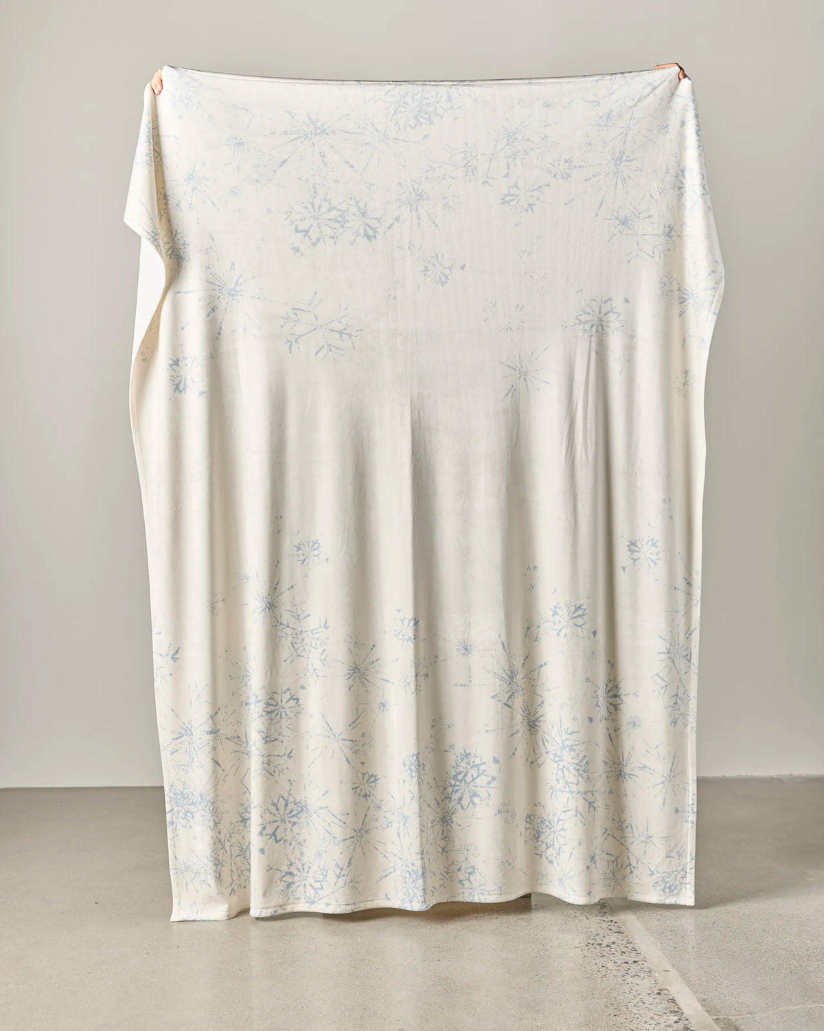 A person holds up Geometry’s “Frosted Windowpanes” throw blanket, featuring a large, off-white design with a subtle pale blue snowflake pattern, against a plain background.