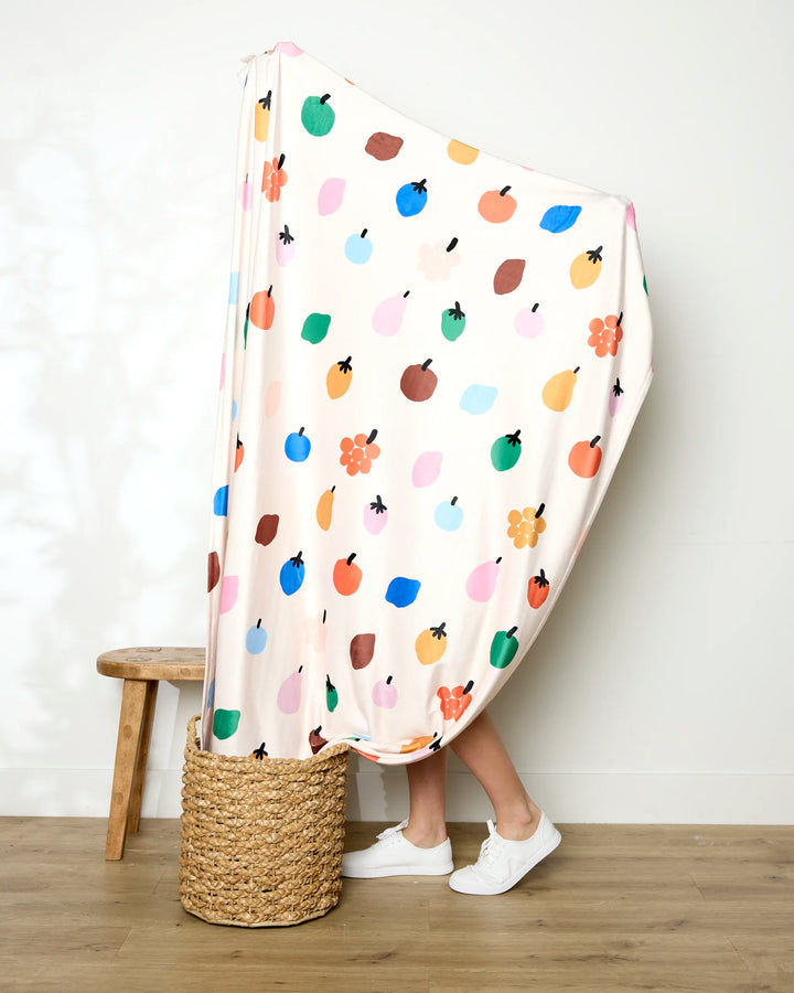 A person stands with their legs visible, wrapped in the colorful Jennifer Bouron Fruit Basket blanket adorned with fruit and flower patterns. They wear white sneakers next to a wooden stool and a woven basket on a wooden floor.