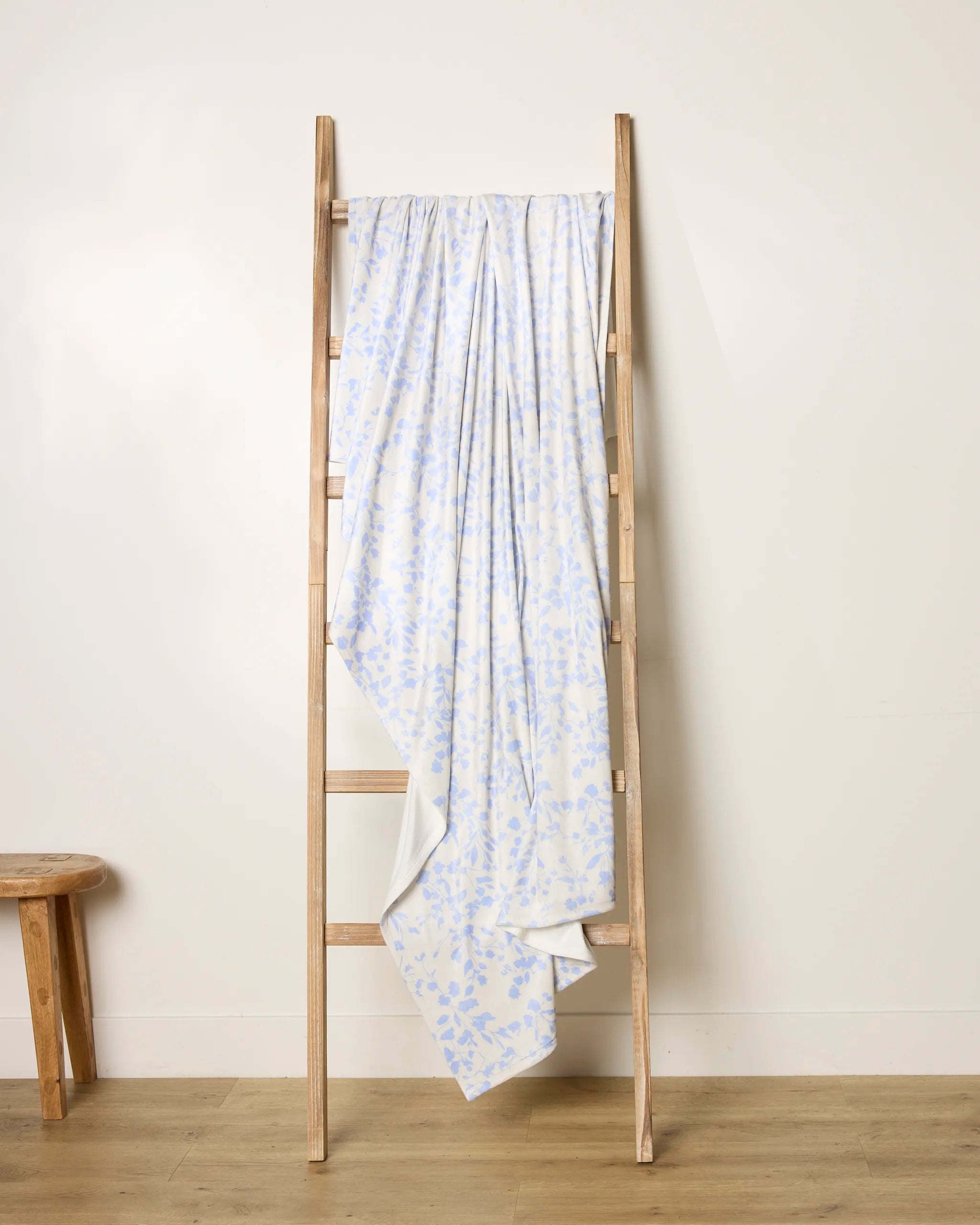 A light wooden Glowgarden ladder by Geometry leans against a white wall, draped with a soft pale blue and white throw; to the left, a small wooden stool rests on a light wood floor.