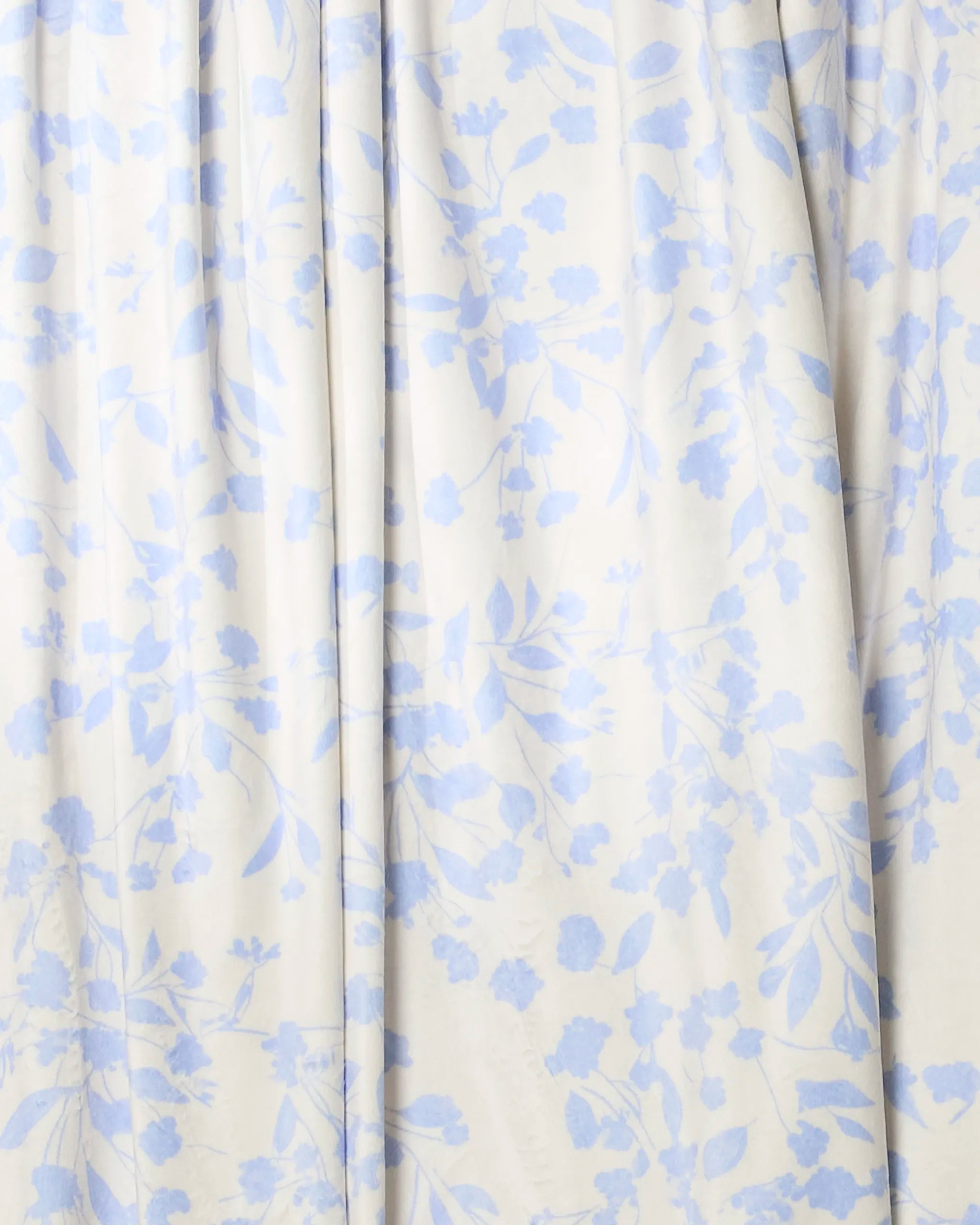 A close-up of Glowgarden by Geometry, a soft throw blanket with pale fabric, features a light blue floral pattern of leaves and small flowers. The gently gathered material creates subtle folds and shadows.