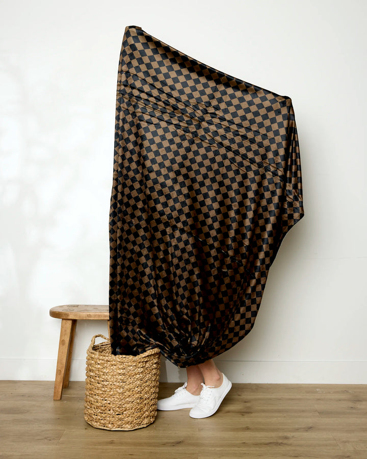 A person stands on a wooden floor with only their lower legs and white sneakers visible, draped in Geometry’s Gridlock large black and brown checkered soft throw blanket beside a woven basket and wooden stool against a plain white wall.