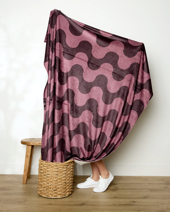 A person stands behind the Geometry Groove With Me blanket, which features a pink and purple wavy pattern and covers them except for their white sneakers and lower legs. A woven basket and wooden stool sit on the wooden floor beside this soft throw.