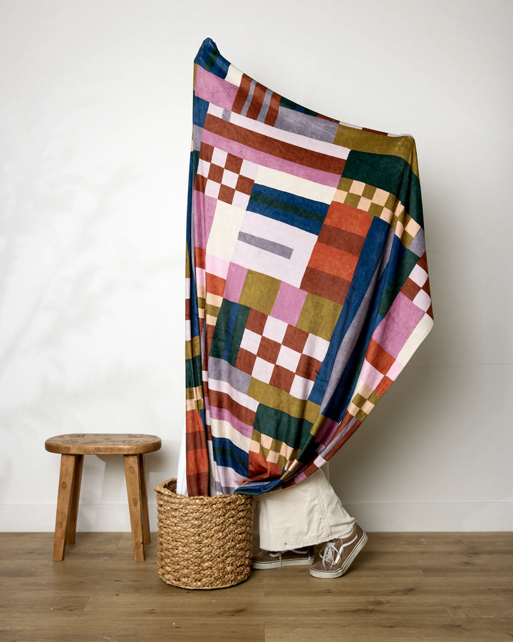 A person stands behind the Lisa Nelson Heirloom Quilt, a colorful geometric-patterned throw. Only their shoes and pant hem show below. Nearby, a small wooden stool sits on the wooden floor.