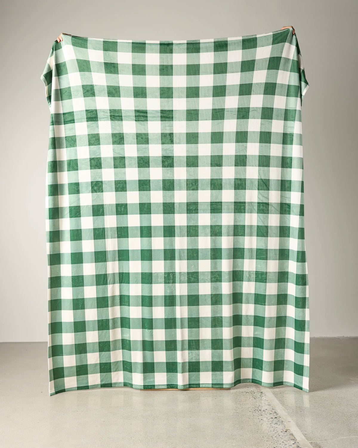 A Geometry Holly Check soft throw blanket, featuring a large green and white checkered pattern, is being held up against a plain wall, with only the fingers of two people visible at the top corners.