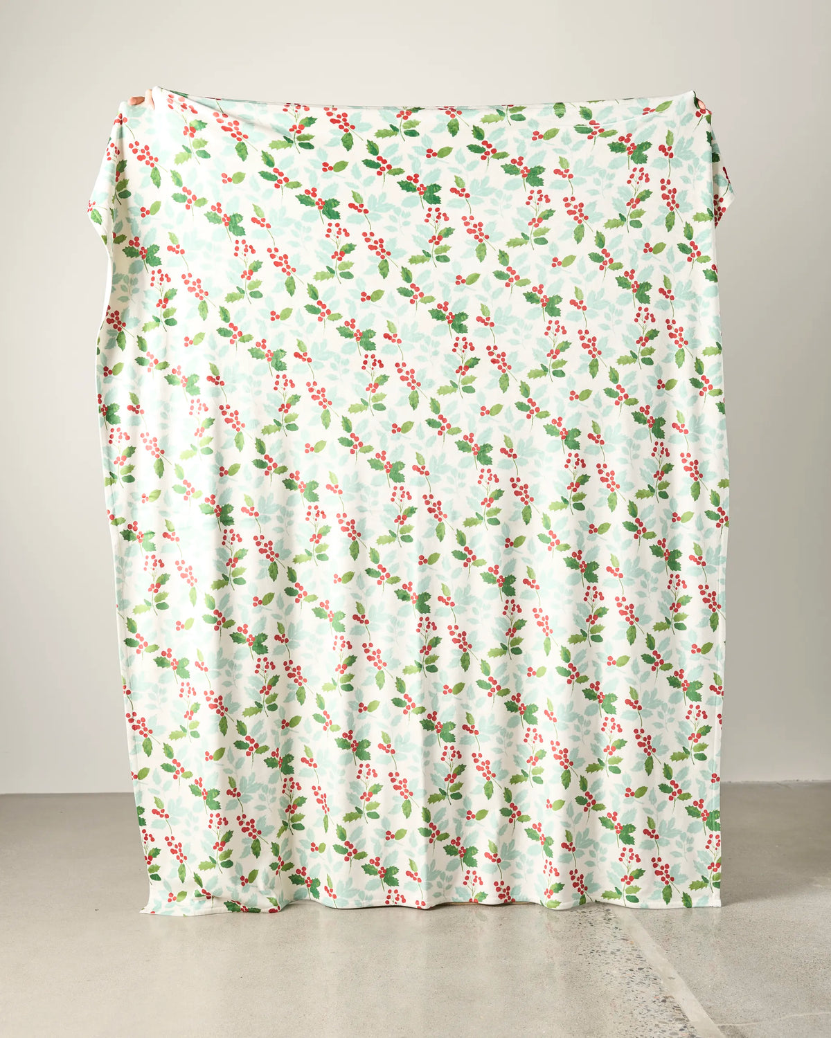 The Geometry Holly Lane Butter Blanket, featuring a festive green and red holly pattern, is draped vertically against a plain, light-colored wall and floor.