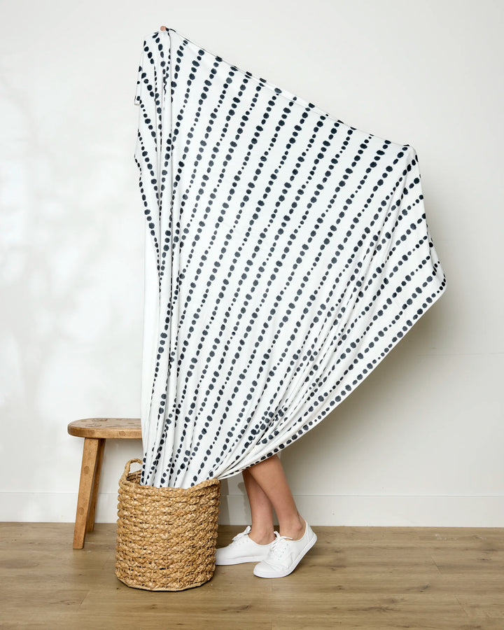 A person stands in a woven basket, holding up the Geometry Ink Dots throw blanket with diagonal black dotted lines; only their legs and white sneakers are visible. A wooden stool sits nearby against a plain wall.