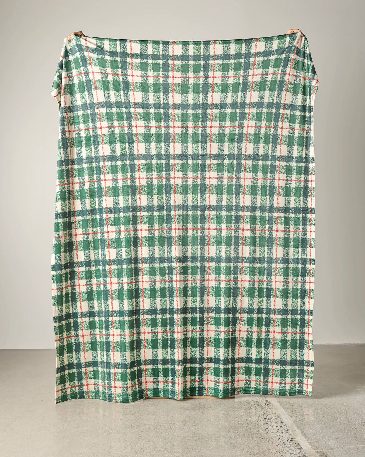 A large Juniper Plaid throw blanket by Geometry, featuring a green, white, and red plaid pattern, is held up against a plain, light-colored wall and floor background. This soft blanket blends cozy comfort with classic style—perfect for adding warmth to any space.