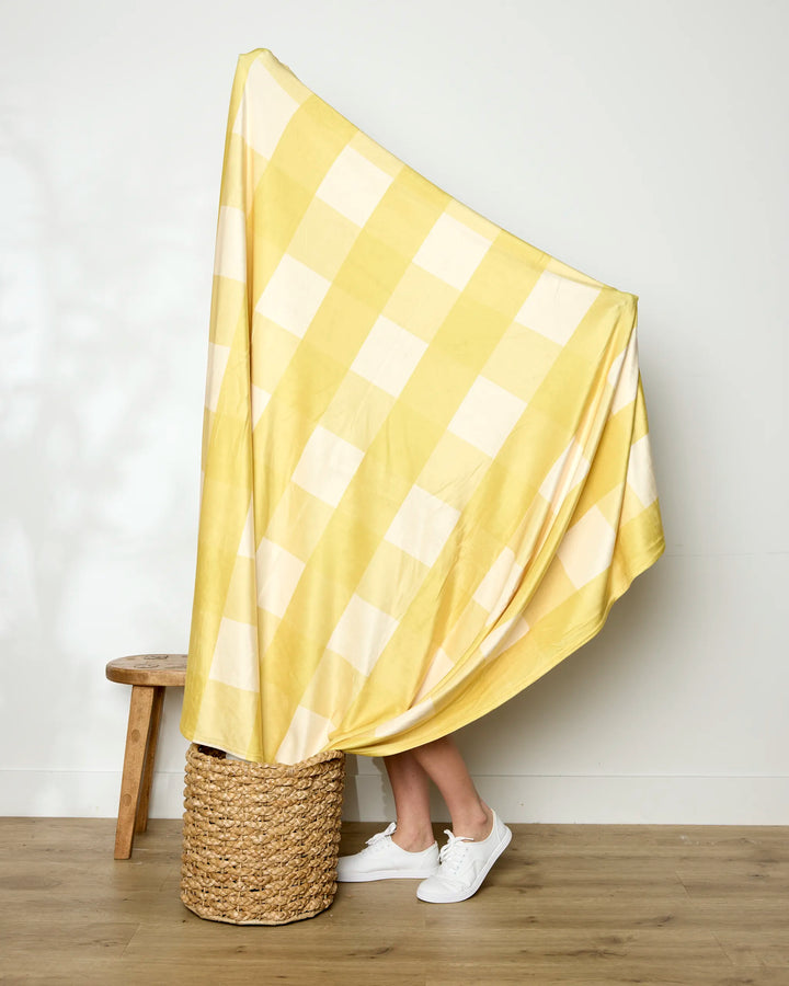 A person stands on a woven basket, mostly hidden behind Geometry's Lemonade Gingham blanket, with only their lower legs and white sneakers visible beside a wooden stool.