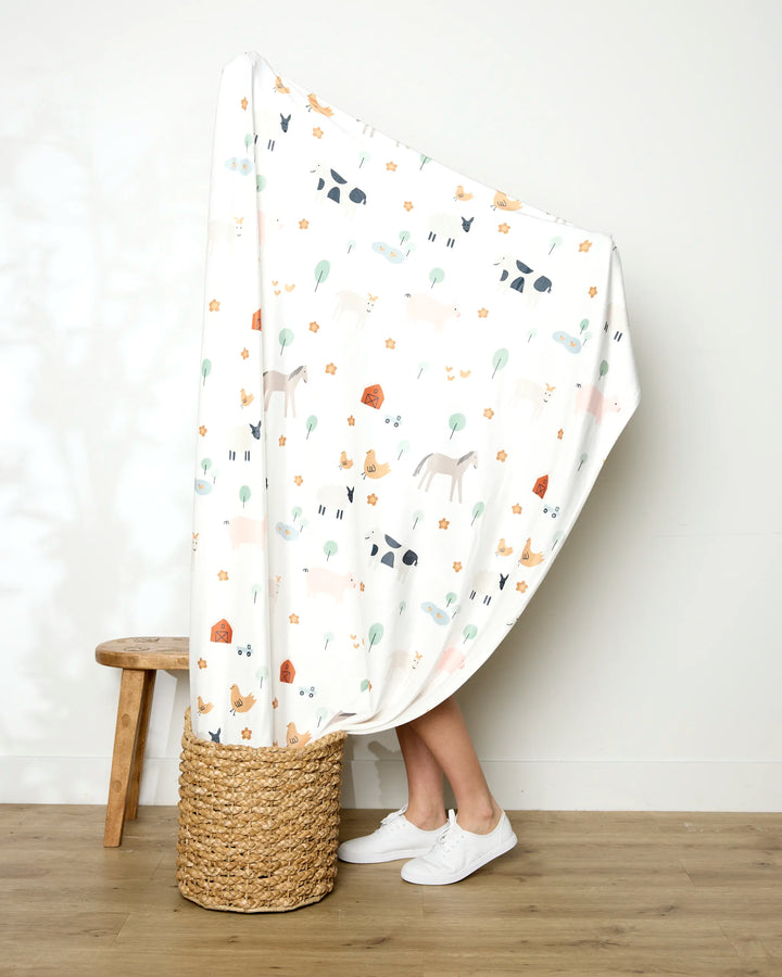 A person stands in a woven basket, wrapped mostly in the Coit Creative Light Farm Animals blanket with animal and plant patterns. Only their lower legs and white shoes are visible. A wooden stool rests nearby against a plain wall and wood floor.