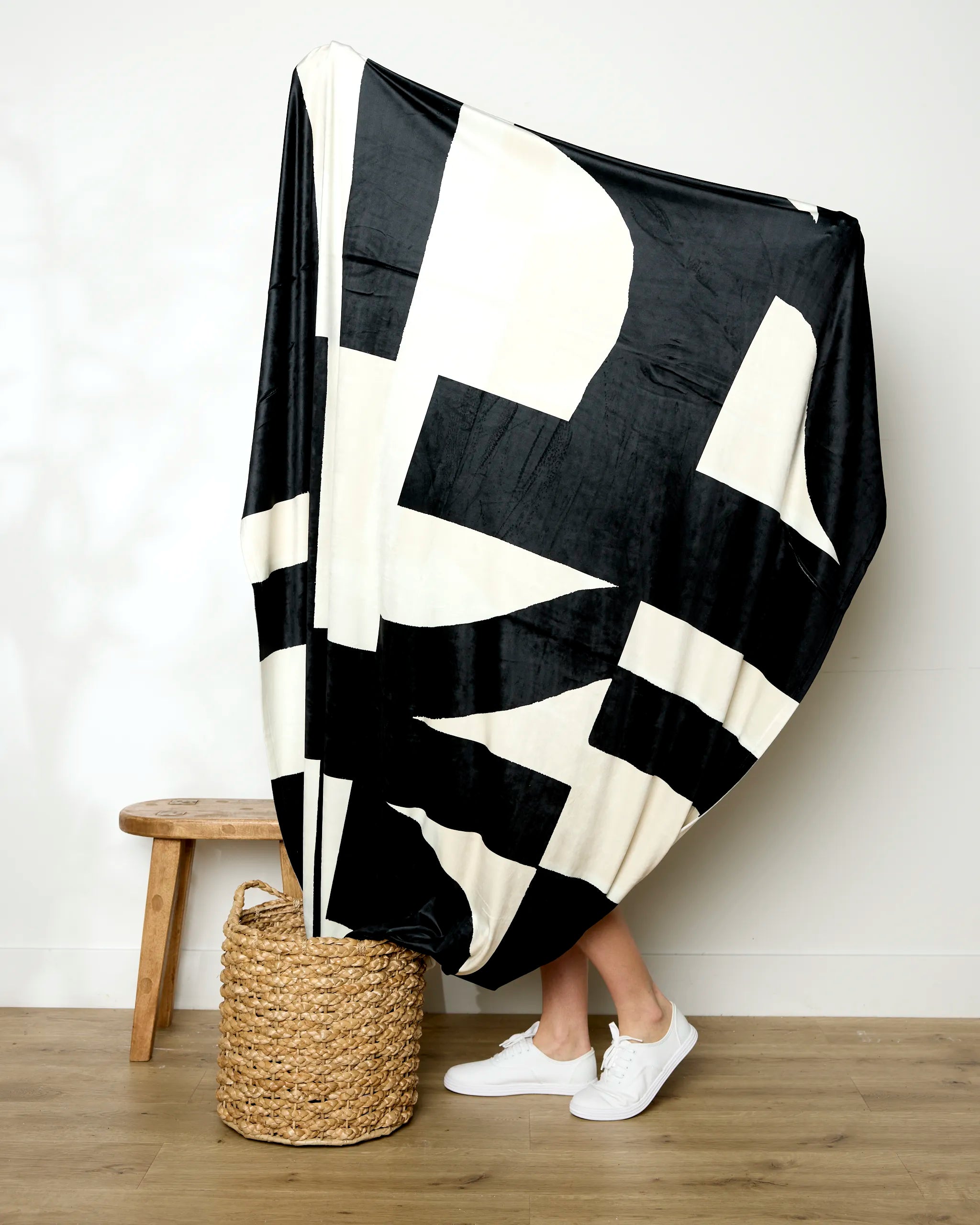 A person stands partially hidden behind a large black-and-white Lined Square blanket from Geometry, with only their lower legs and white sneakers visible. A woven basket and wooden stool add warmth to the cozy wooden-floored scene.
