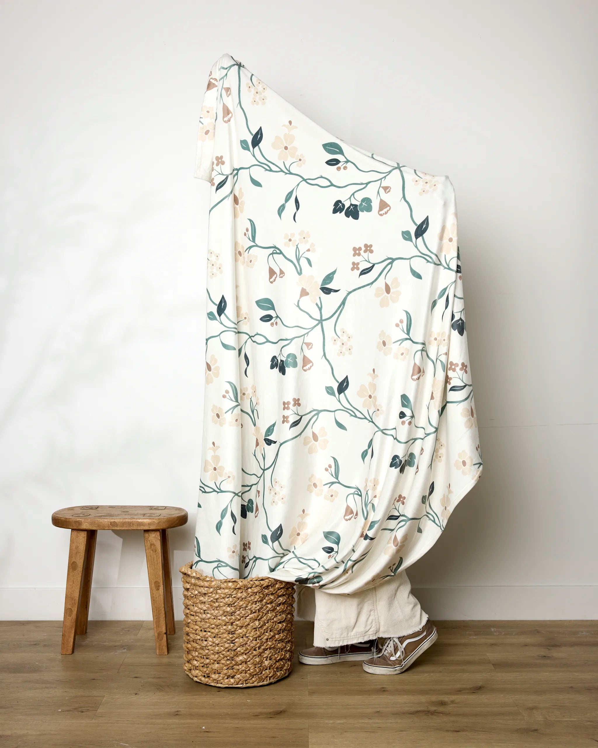 A person stands behind the Danelys Sidron "Lunaria in Alabaster" recycled blanket, featuring a cream botanical pattern. Only their shoes and pant legs are visible, with a wooden stool to the left and a woven basket in front of them.
